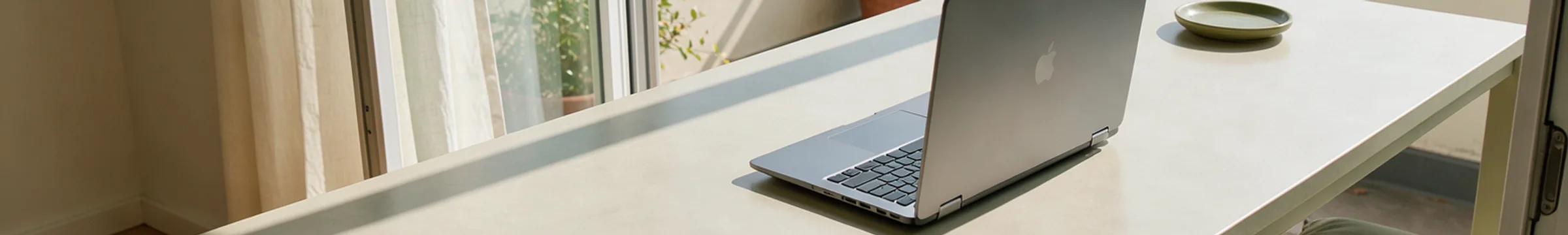 An open personal laptop seen from behind on a bright apartment balcony, with a curtain shadow across the table.